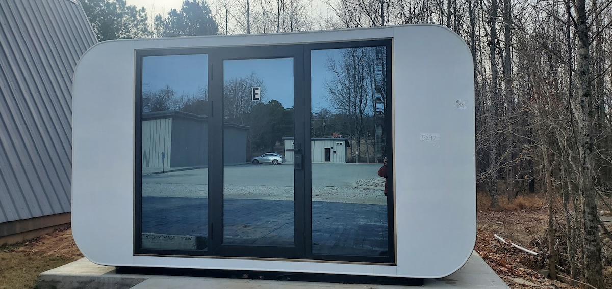 The exterior of the tiny cube is highlighted by large, dark-framed glass doors that reflect the surrounding trees. The minimalist structure features a rounded top and is positioned on a concrete base, with a gravel driveway visible in the background.