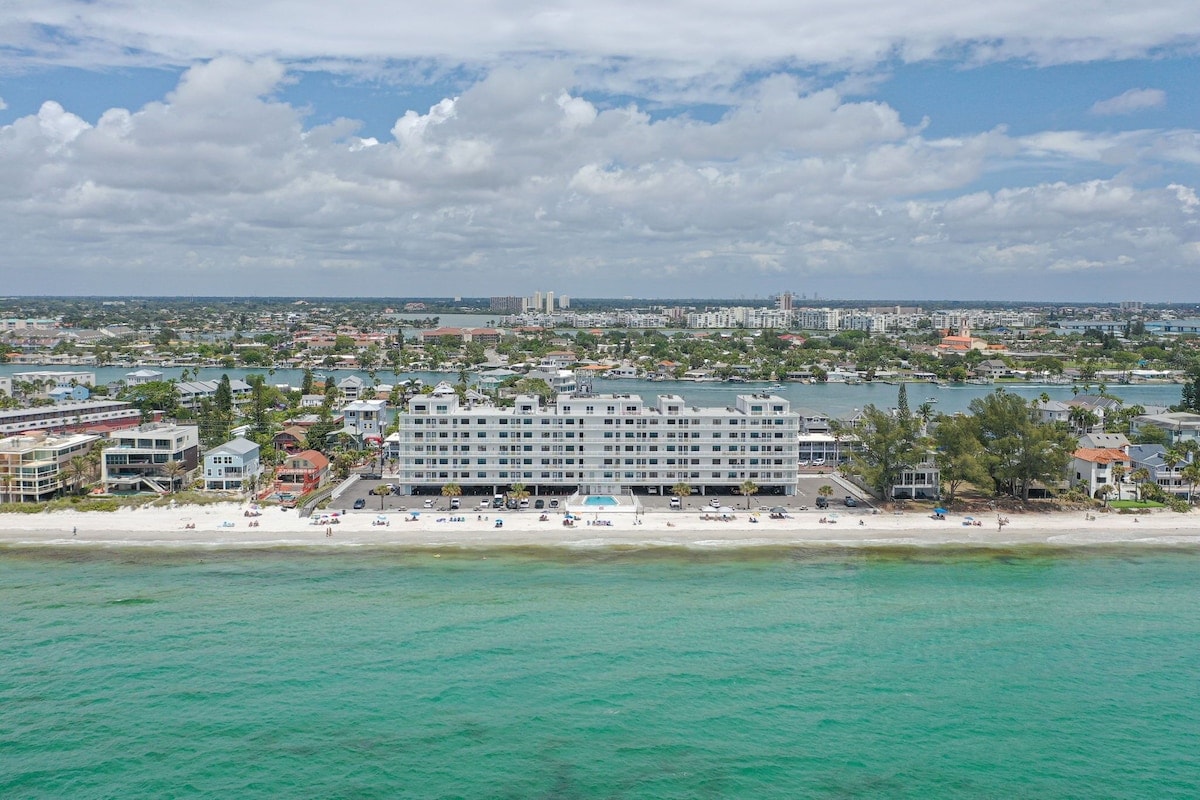 An aerial view captures a multi-story beachfront condominium situated along the Gulf of Mexico. Sandy shores line the base, while the turquoise ocean extends towards the horizon. Nearby buildings and vegetation are visible, providing context to the serene coastal environment.