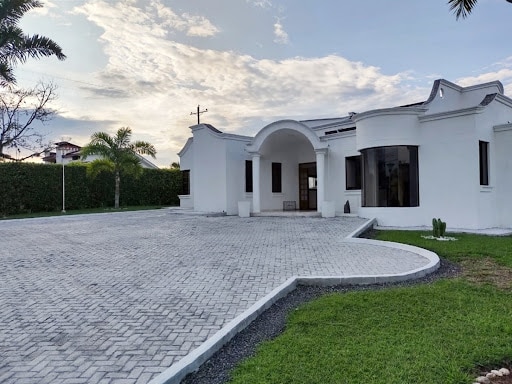 A modern, single-story home features an arched entrance and large windows that provide abundant natural light. The surrounding landscape includes well-maintained grass areas and palm trees, while a stone-paved driveway enhances the outdoor appeal.