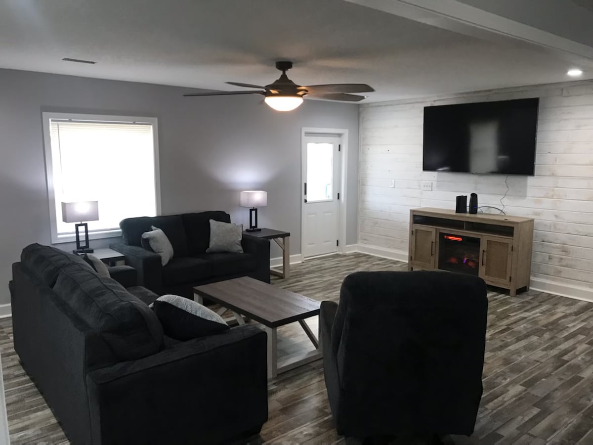 A living area features a comfortable arrangement of dark-colored sofas and armchairs. A wooden coffee table is centered among the seating. A television is mounted on the wall above a console with a warming feature. Natural light enters through a window with sheer coverings.