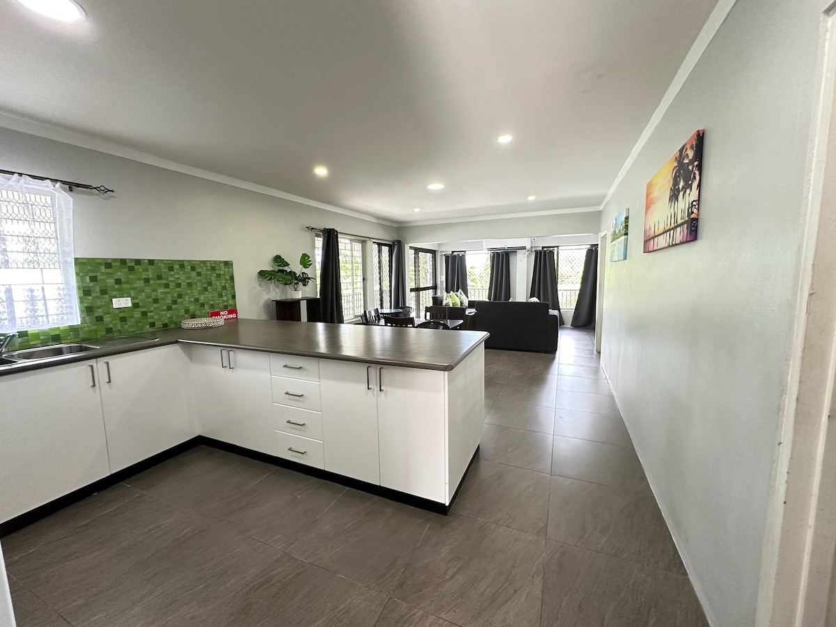 An open-plan kitchen is shown, featuring white cabinetry and a dark countertop. A green tiled backsplash adds color. Natural light fills the living area, where dark curtains frame the windows. The space is designed for both cooking and socializing, enhancing the apartment's inviting layout.