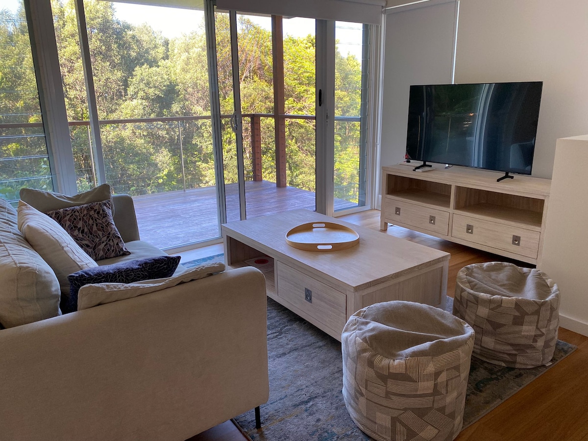 A spacious living area features a comfortable seating arrangement with a light-colored sofa and accent cushions. A wooden coffee table sits in the center, accompanied by two poufs. Large glass doors lead to a private balcony surrounded by lush greenery.