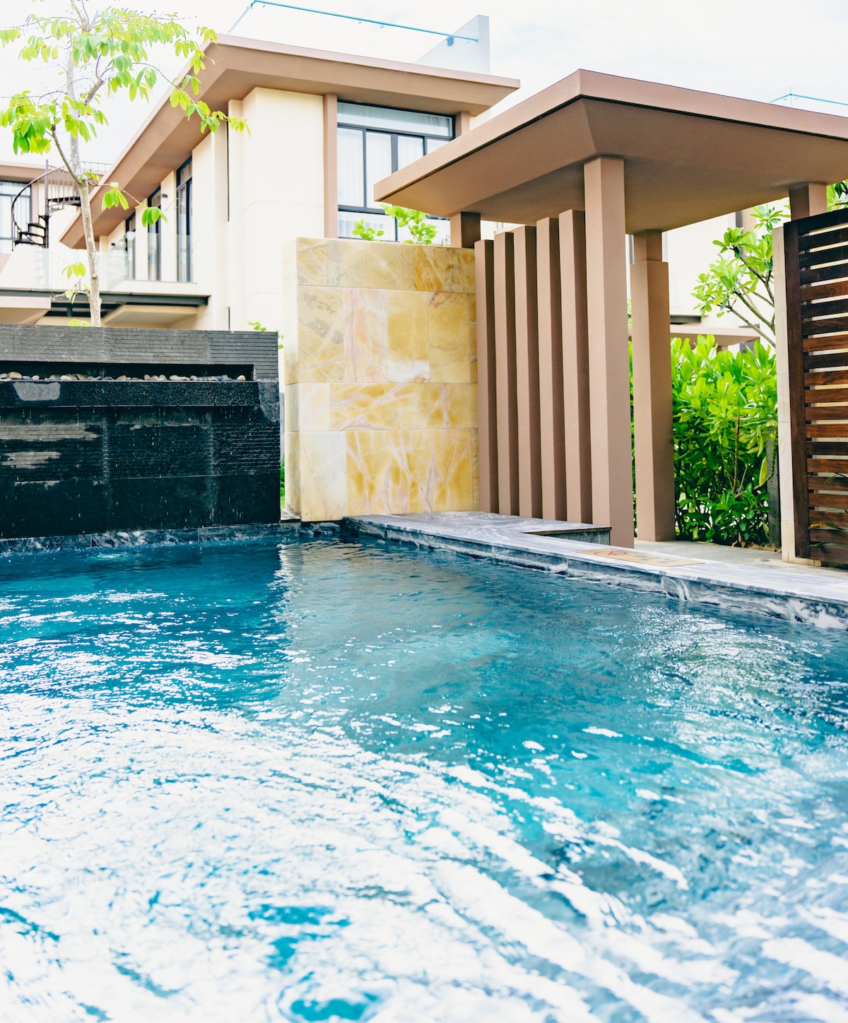 An infinity pool is featured with clear blue water, adjacent to a modern villa structure. The pool's edge extends seamlessly, allowing for picturesque reflections. Surrounding greenery complements the serene atmosphere, while the villa's architectural elements provide a contemporary backdrop.