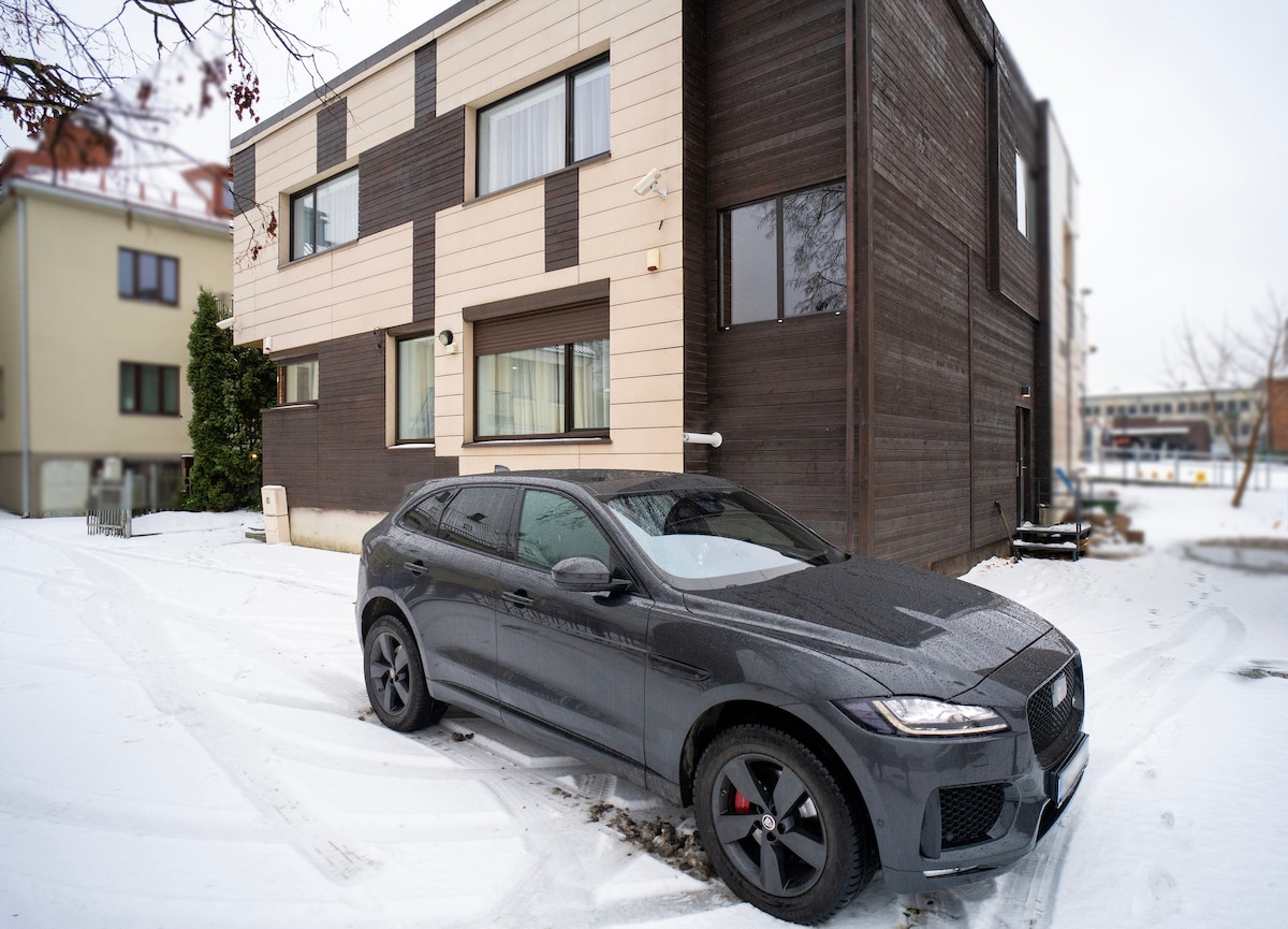 A modern two-story building features a combination of dark wood and light-colored panels. In the foreground, a sleek black vehicle is parked on a snow-covered driveway, highlighting the winter setting. The flat roof and large windows give the structure a contemporary appearance.