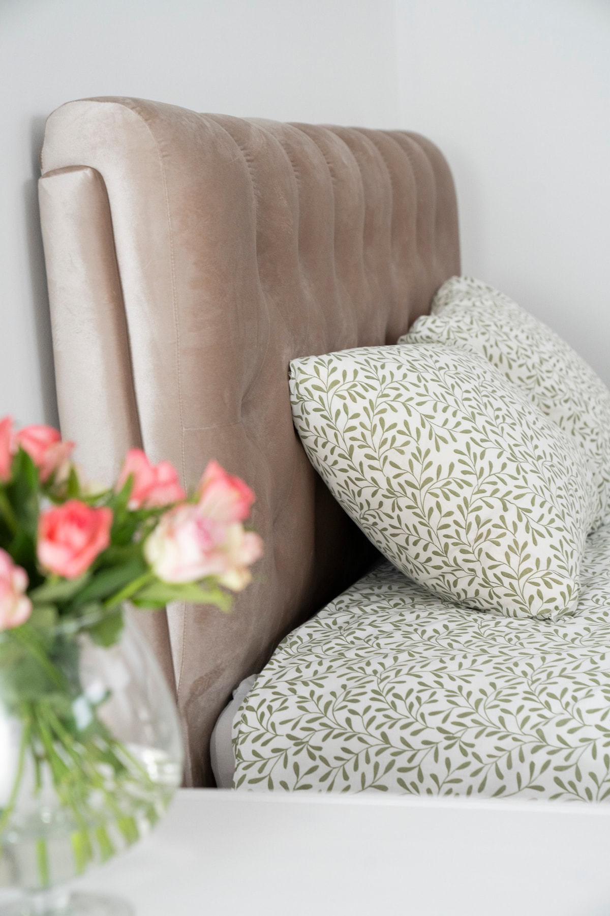 A plush bed with a tufted headboard is featured against a neutral wall. Fluffy pillows with a delicate leaf pattern complement the bed. A clear glass vase filled with fresh flowers adds a touch of color to the scene.