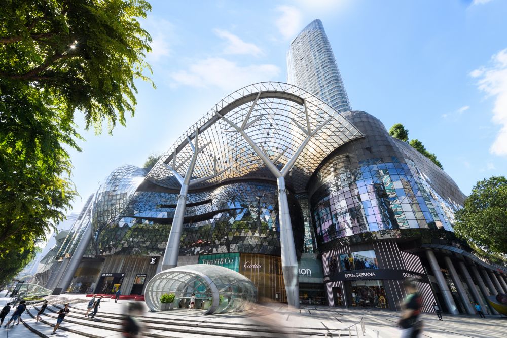 A modern shopping complex features an intricate, glass-covered design with flowing curves and numerous reflective surfaces. Lush greenery is integrated into the architecture, enhancing the urban landscape. Pedestrians are visible, adding life to the scene around the vibrant retail space.