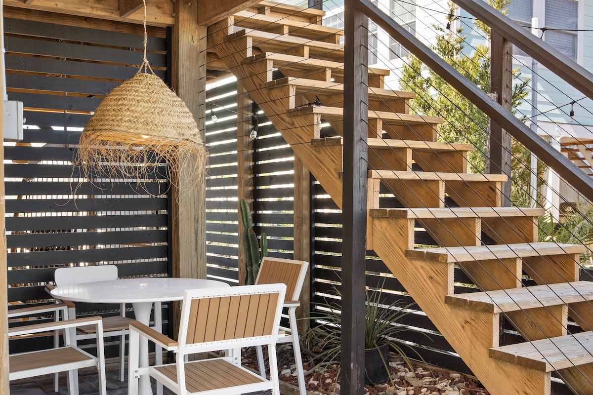 A cozy outdoor seating area is located under a wooden deck. A round table with four white chairs is surrounded by natural wood elements. Stairs ascend to the upper level, framed by a modern wooden slatted wall, offering a blend of shade and openness.