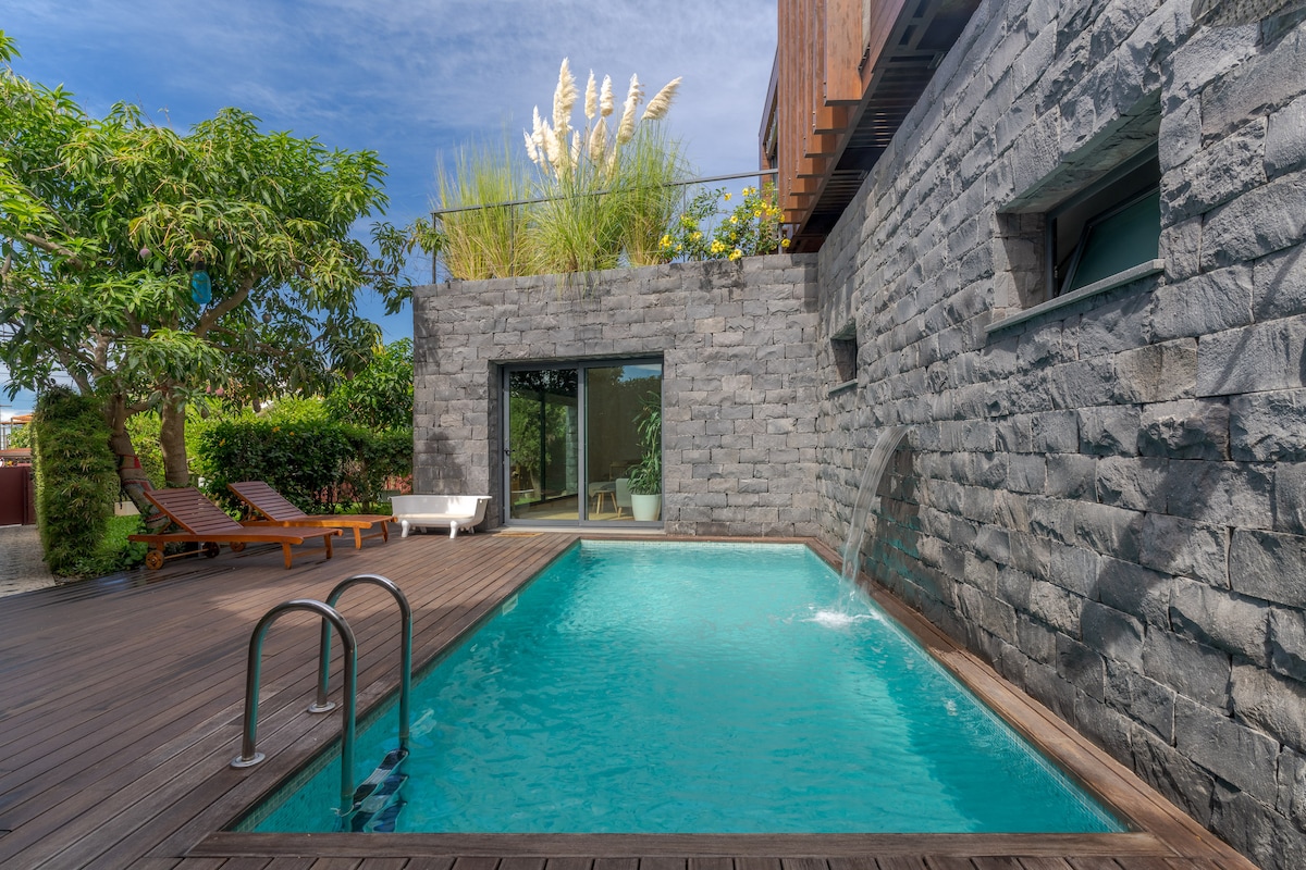 A modern stone-walled pool area features a turquoise water surface with a gentle waterfall. Sun loungers are positioned neatly alongside the pool, surrounded by lush greenery and ornamental grasses, creating a serene outdoor environment.