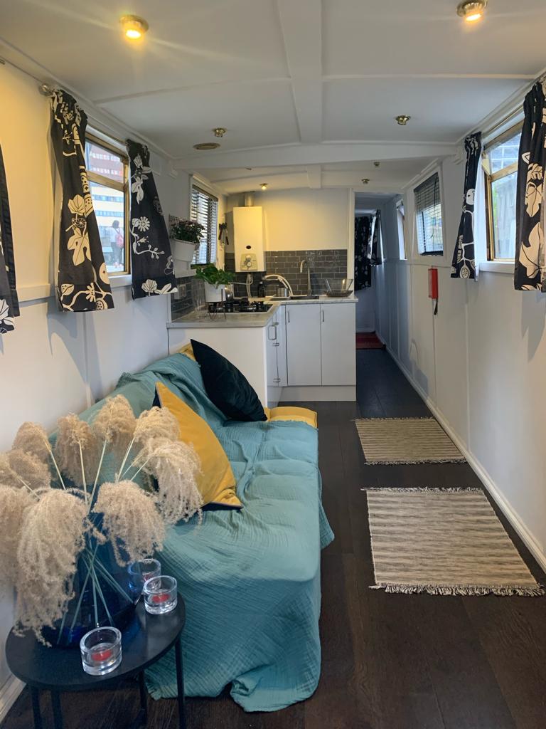 An open-plan living area is featured with a cozy seating arrangement accented by colorful cushions. Dark-colored curtains frame the windows, while a small kitchen area equipped with a gas hob and sink is visible. Decorative grasses in a vase add a touch of warmth to the space.