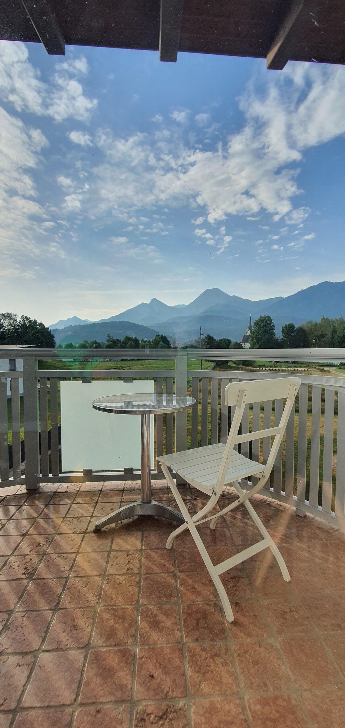 A private balcony is visible, featuring a small round table and a white folding chair. The surrounding landscape displays lush greenery, with distant mountains against a backdrop of cloudy blue skies, suggesting a serene outdoor space for relaxation.