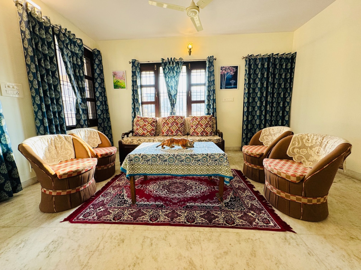 A spacious living area features four comfortable chairs and a traditional sofa arranged around a central table. The floor is adorned with a decorative rug. Tall windows are draped with patterned curtains, allowing natural light to brighten the room.