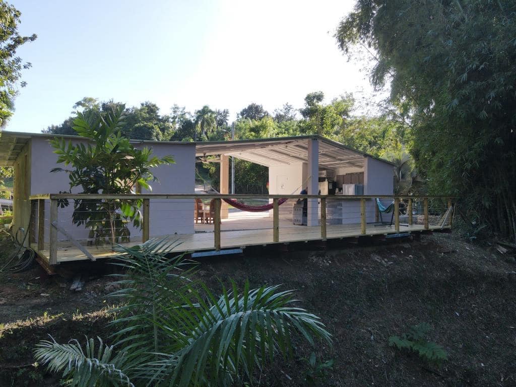 A single-story trailer, designed with an open concept, is surrounded by lush greenery. A spacious deck is visible, featuring a relaxing hammock. The structure's exterior showcases light-colored walls, and large windows allow natural light to fill the interior.