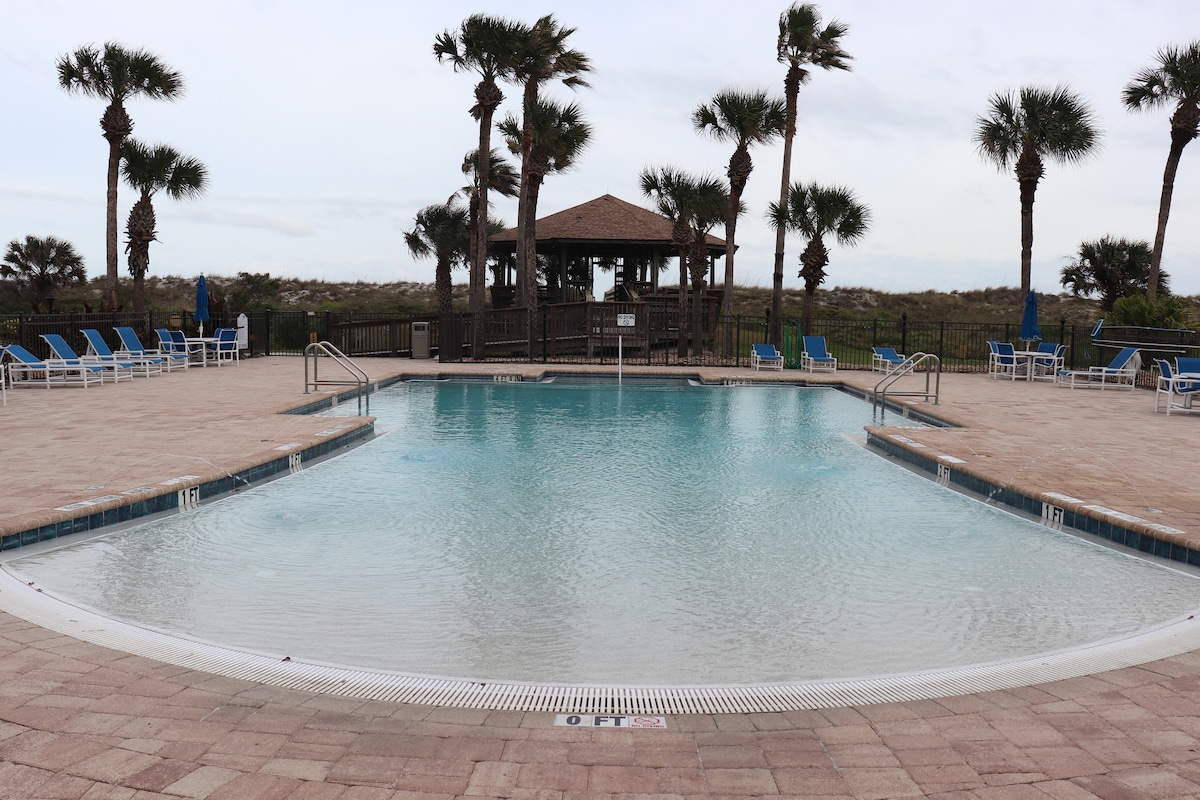 A spacious pool area is surrounded by tiled decking, featuring lounge chairs and a shaded gazebo in the background. Palm trees gently sway in the breeze, enhancing the inviting atmosphere of the outdoor space. The pool appears calm and clear, ready for use.