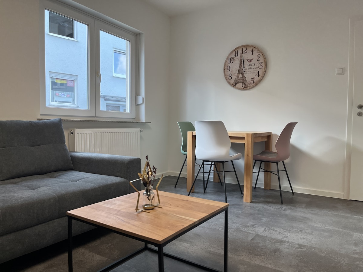 A comfortable living space is featured, showcasing a gray sofa beside a wooden coffee table with decorative elements. A light-colored dining table is accompanied by four chairs in varying colors. Natural light pours in through two windows, brightening the inviting atmosphere.