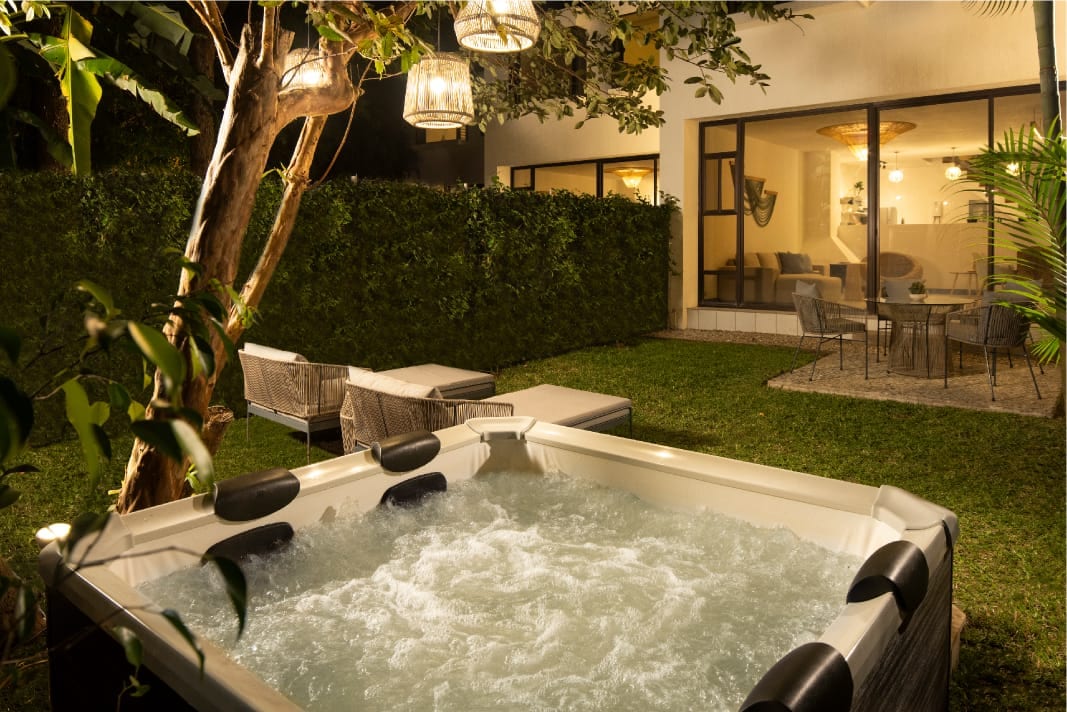 A private jacuzzi bubbles gently in the garden, illuminated by soft outdoor lighting. Comfortable lounge chairs are positioned nearby, surrounded by lush greenery that provides a sense of privacy. Large glass doors reveal a cozy seating area within the villa.