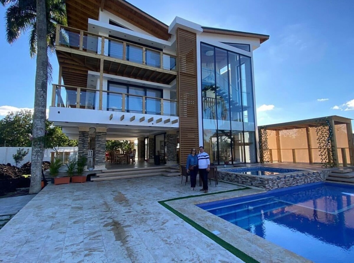 A modern two-story home features expansive glass walls and a spacious terrace that overlooks a swimming pool. Palm trees line the property, enhancing the outdoor space. The structure is illuminated by natural light, creating a serene and open atmosphere.