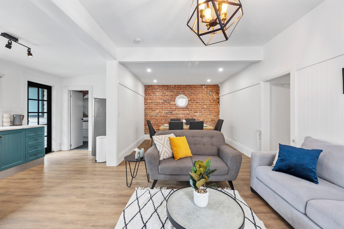 The open living area features light-colored wood flooring and exposed brick walls. A grey sofa and an inviting armchair are arranged around a round coffee table with greenery. The dining space includes a table set for six, illuminated by a stylish pendant light.