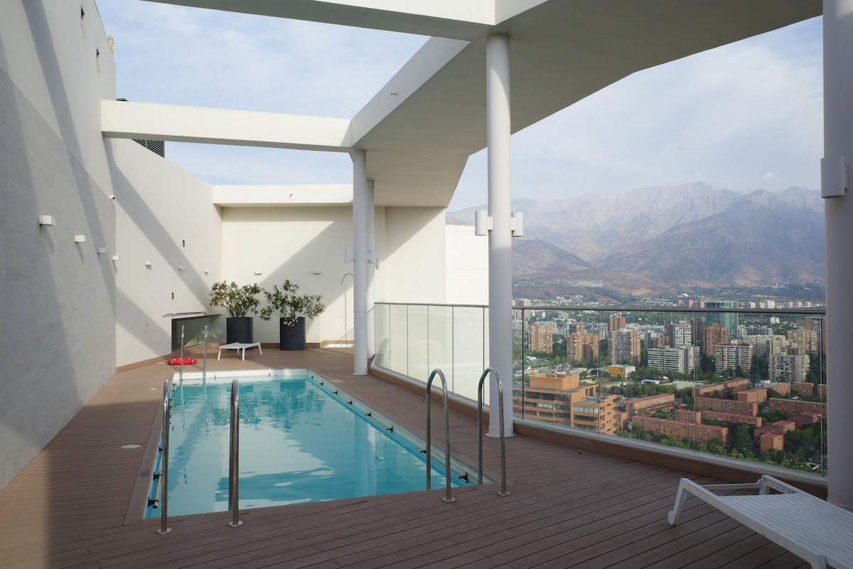 An outdoor rooftop pool is framed by sleek white walls and a modern deck. Panoramic views of the city and the Andes mountains are visible in the background. Sun loungers are placed nearby, creating a spacious area for relaxation.