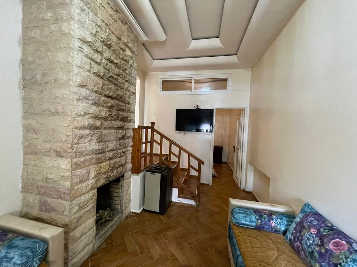 A spacious living area features a stone fireplace as a focal point. Light-colored walls provide a neutral backdrop for the room. A television is mounted on the wall, and a staircase with wooden railing leads to an upper level. Comfortable seating areas are arranged nearby.