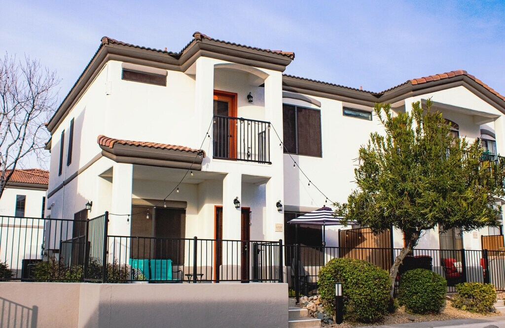 A two-story building with a modern design is framed by a wrought iron fence. Balconies are positioned on both levels, featuring sliding doors. A shaded area with outdoor seating is visible on the ground floor, along with neatly trimmed shrubs and a small tree in the foreground.