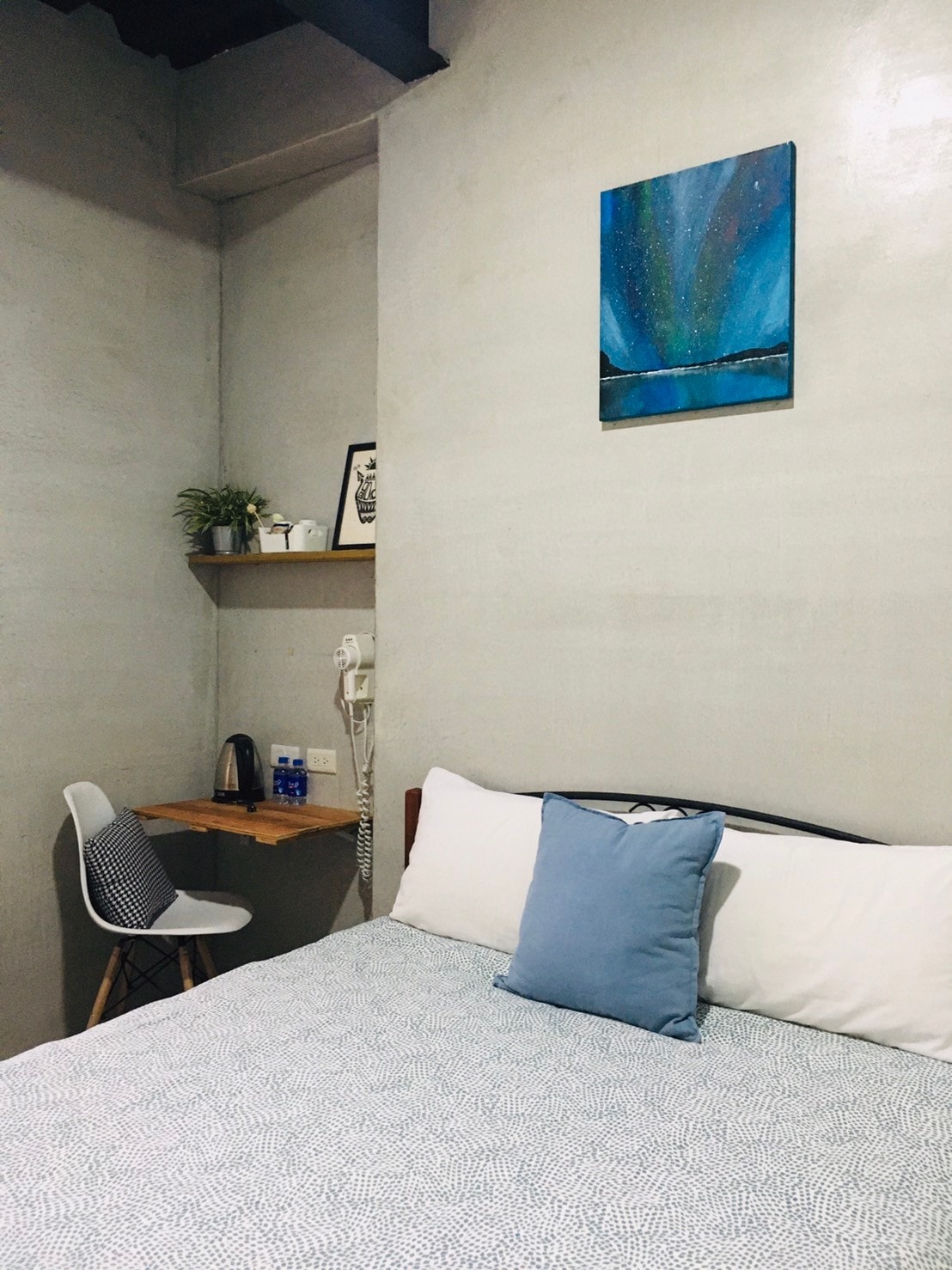 A cozy bedroom features a spacious bed with soft white and blue bedding. A simple desk with a chair is positioned against the wall, accompanied by a small plant and framed art. Light blue and gray tones create a calm environment.