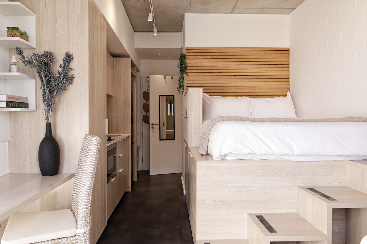 A modern studio apartment features a raised sleeping area with a spacious bed, complemented by light wood finishes. A small kitchenette and dining area are visible, with a textured wall adding subtle detail. Large windows invite natural light, enhancing the bright and airy ambiance.