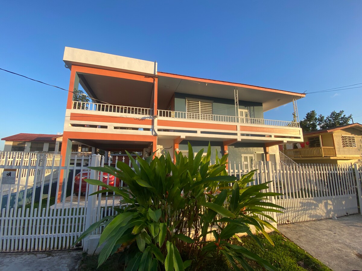 A two-story building features a modern design with an inviting color palette. The exterior showcases a spacious porch area and a well-maintained garden with lush greenery. A white picket fence surrounds the property, offering privacy, while numerous windows allow ample natural light to flow into the interior.