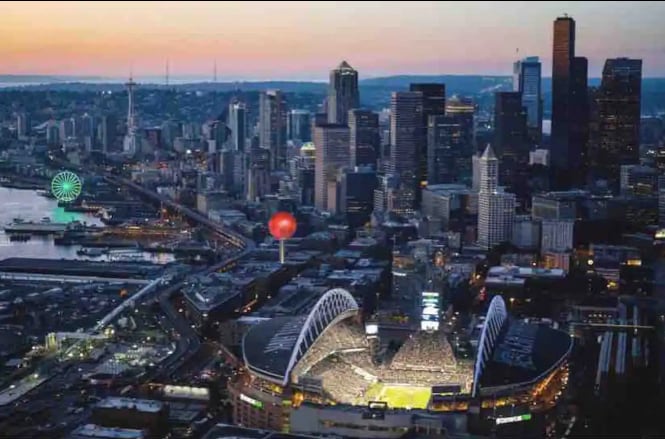 Aerial view of downtown Seattle features various skyscrapers and the iconic Space Needle against a twilight sky. Lumen Field is prominently visible, illuminated during an event. The waterfront includes a Ferris wheel, adding to the vibrant cityscape. The atmosphere is alive with urban energy.