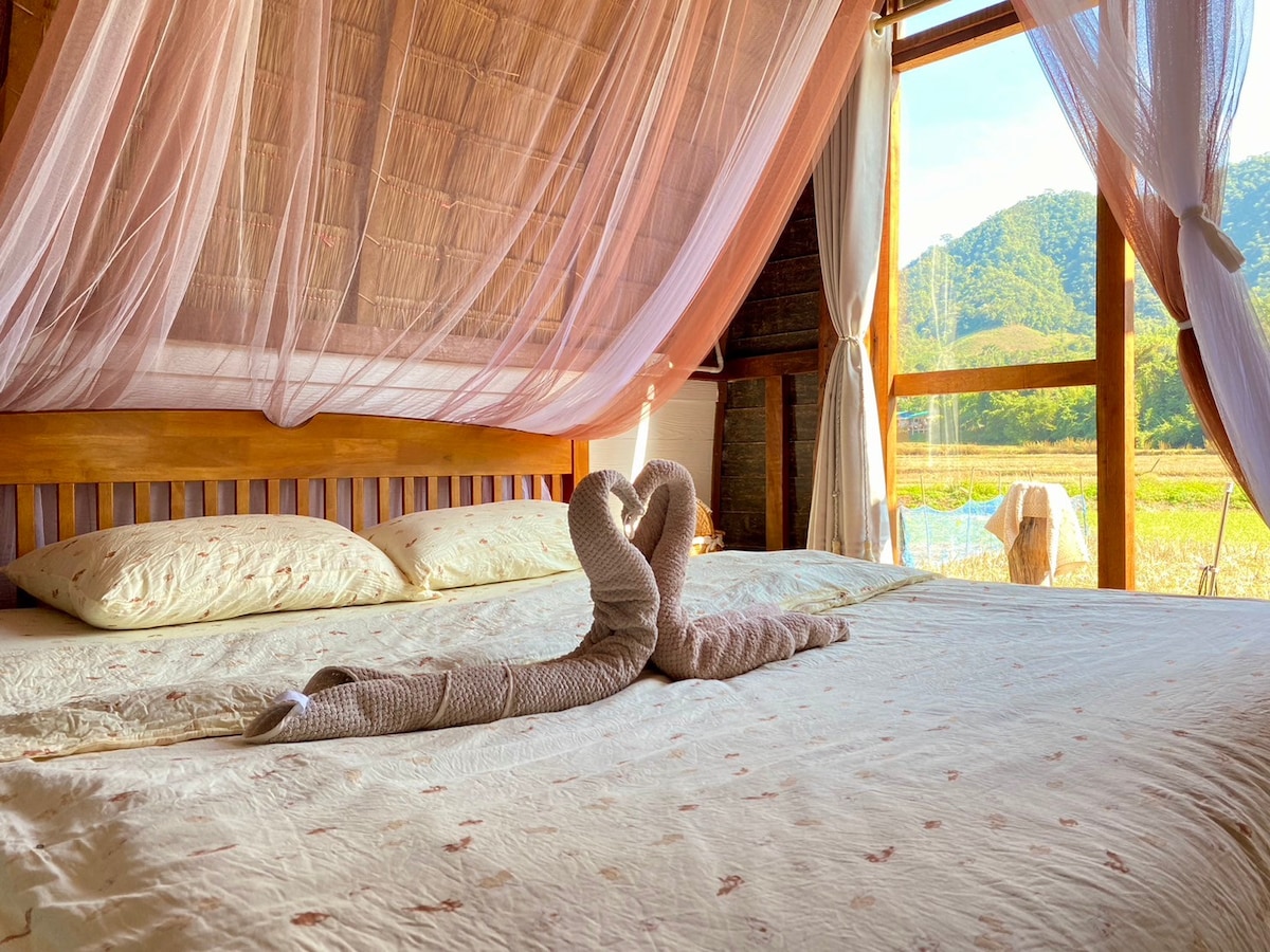 A bed draped with soft, flowing curtains invites relaxation, complemented by a pair of intricately folded towels arranged in a heart shape. Large windows frame a serene view of green hills and a bright sky, enhancing the restful atmosphere of the space.