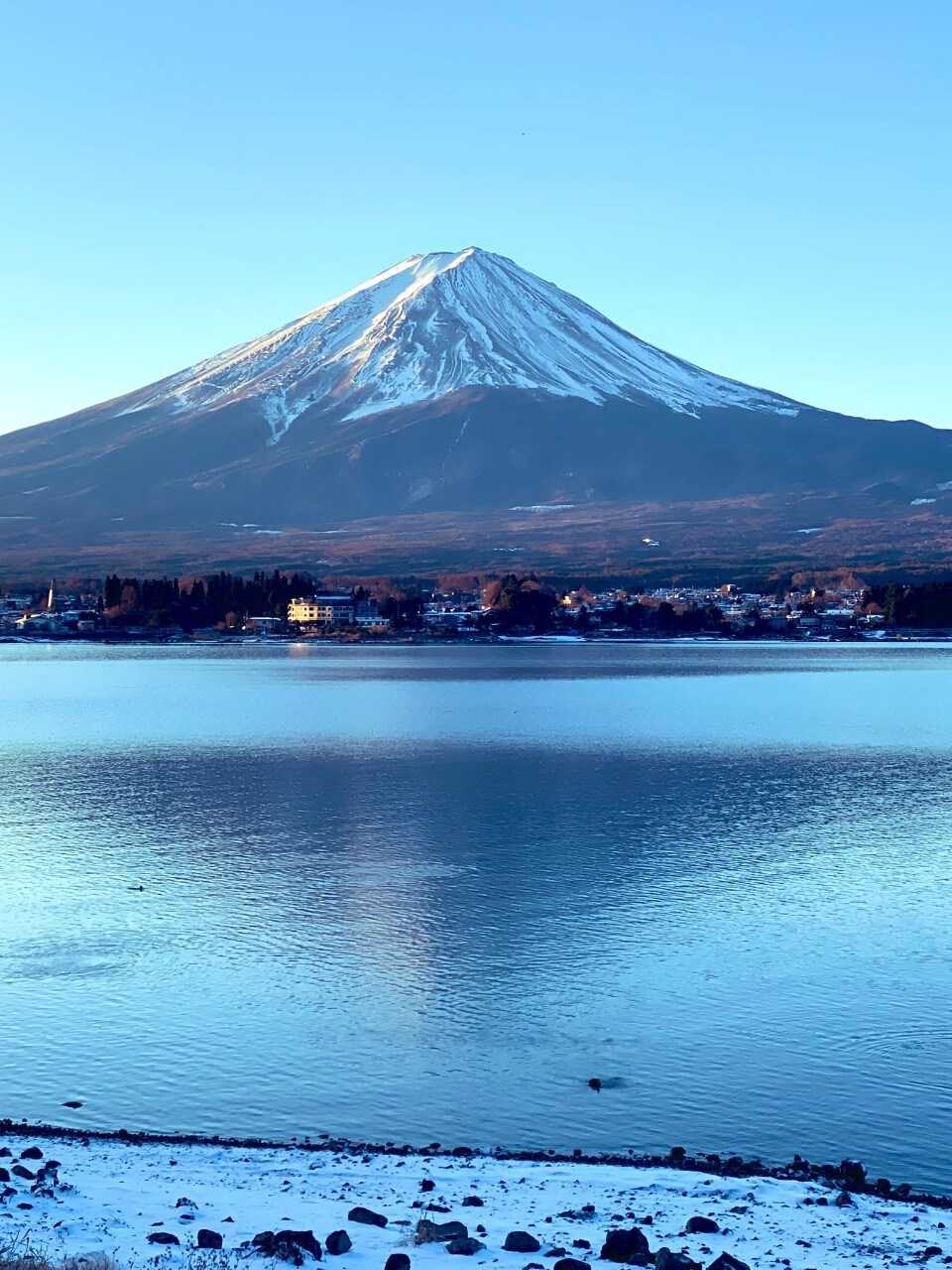 A clear view of Mt. Fuji rises majestically above a calm lake, with snow-capped peaks reflecting in the water. The shoreline features a small settlement, positioned amidst the serene landscape.