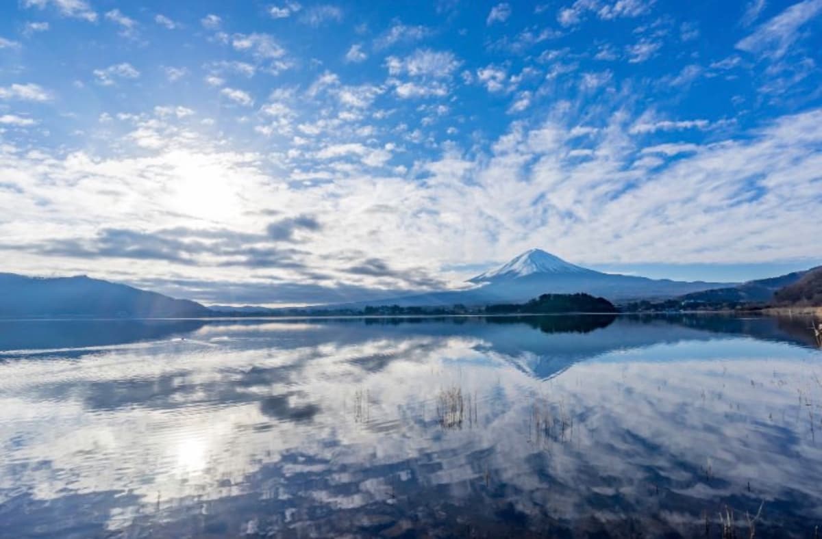 A serene landscape of Lake Kawaguchi is depicted, with a clear reflection of Mt. Fuji and the sky on the water's surface. Soft clouds drift across, while the sunlight illuminates the area, creating a peaceful atmosphere in the early morning light.