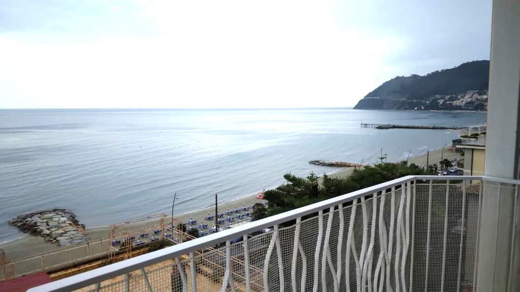 A balcony overlooking a tranquil sea showcases gentle waves lapping at the shore. A distant coastline is visible, framed by a lush hillside. Beach loungers line the sandy beach below, inviting relaxation amidst the serene atmosphere.