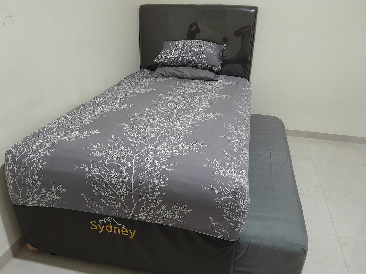 A spacious bed is presented with grey bedding featuring a delicate floral pattern. A second mattress is positioned beside it, enhancing the sleeping arrangement. The light-colored tiled floor and plain wall contribute to a simple and functional atmosphere.