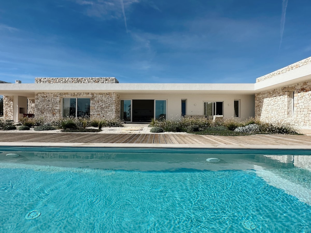 A contemporary villa features a sleek design with a spacious outdoor area. An inviting infinity pool reflects the blue sky, while large glass doors provide views into the open living space. Surrounding greenery includes low-growing plants and natural stone accents.