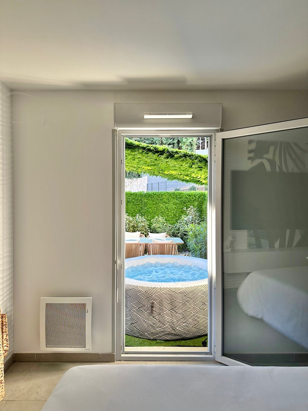 A bright bedroom is shown, featuring a glass door that opens to a private garden area. A jacuzzi is visible outside, surrounded by greenery and a dining setup. The space provides a serene view, enhancing the overall ambiance of the room.
