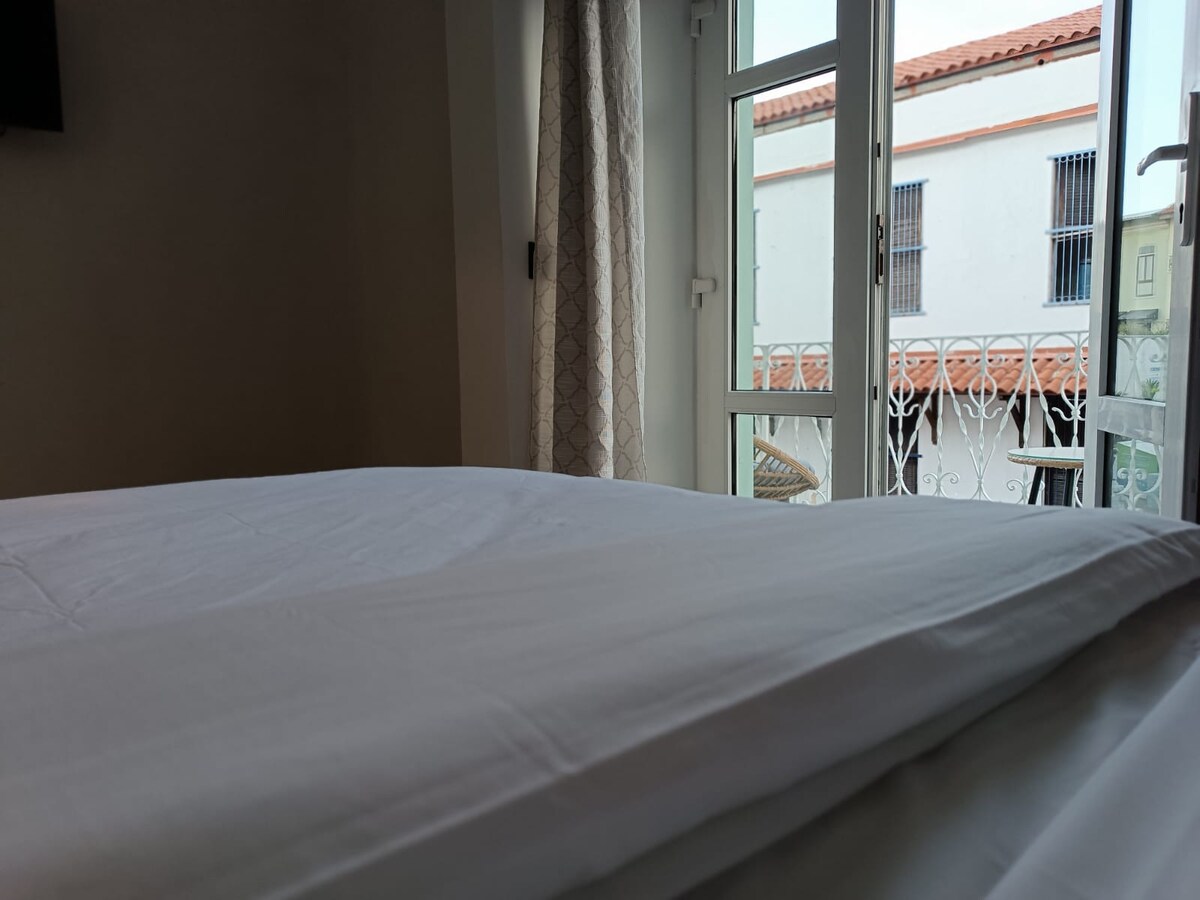 An inviting sleeping area with rumpled white linens is displayed, showcasing a cozy bed positioned near a window. Natural light enters through the window, which is adorned with light-colored curtains, revealing glimpses of a neighboring building's architecture.