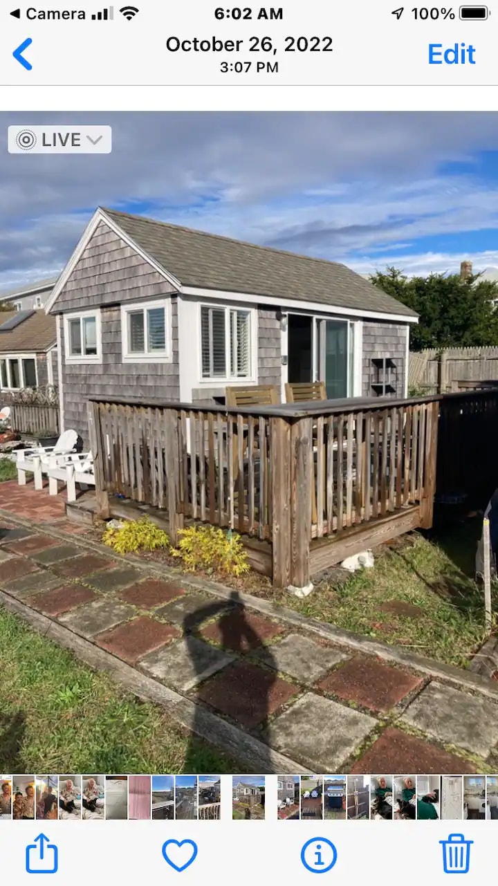 A charming cottage exterior is presented, featuring a well-maintained wooden deck surrounded by a wooden railing. Two white chairs are positioned on the deck, inviting relaxation. The cottage's light-colored siding contrasts with the blue sky and hints of greenery nearby.