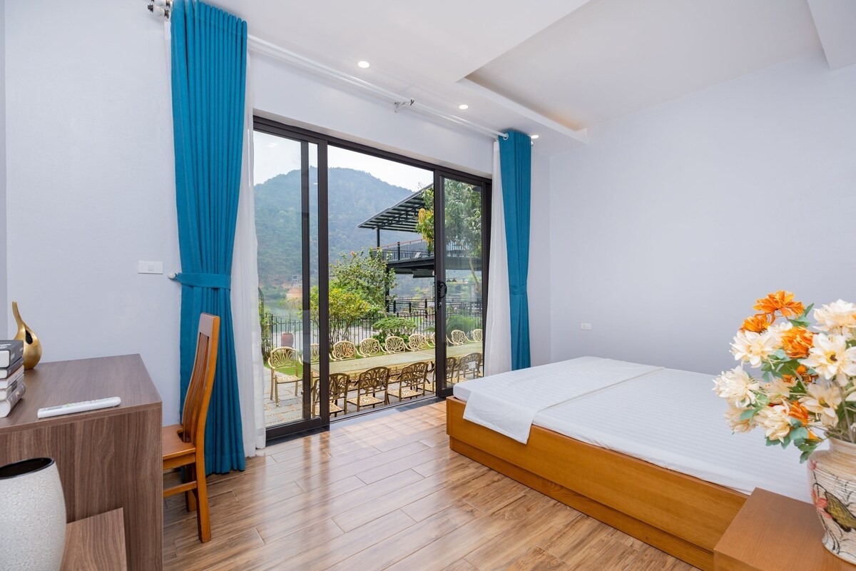 A light-filled room features a spacious bed with a simple white bedspread. Floor-to-ceiling glass doors open to a scenic view of the mountains, while a wooden desk and chair provide a functional workspace. Soft blue curtains add a touch of color to the neutral walls.