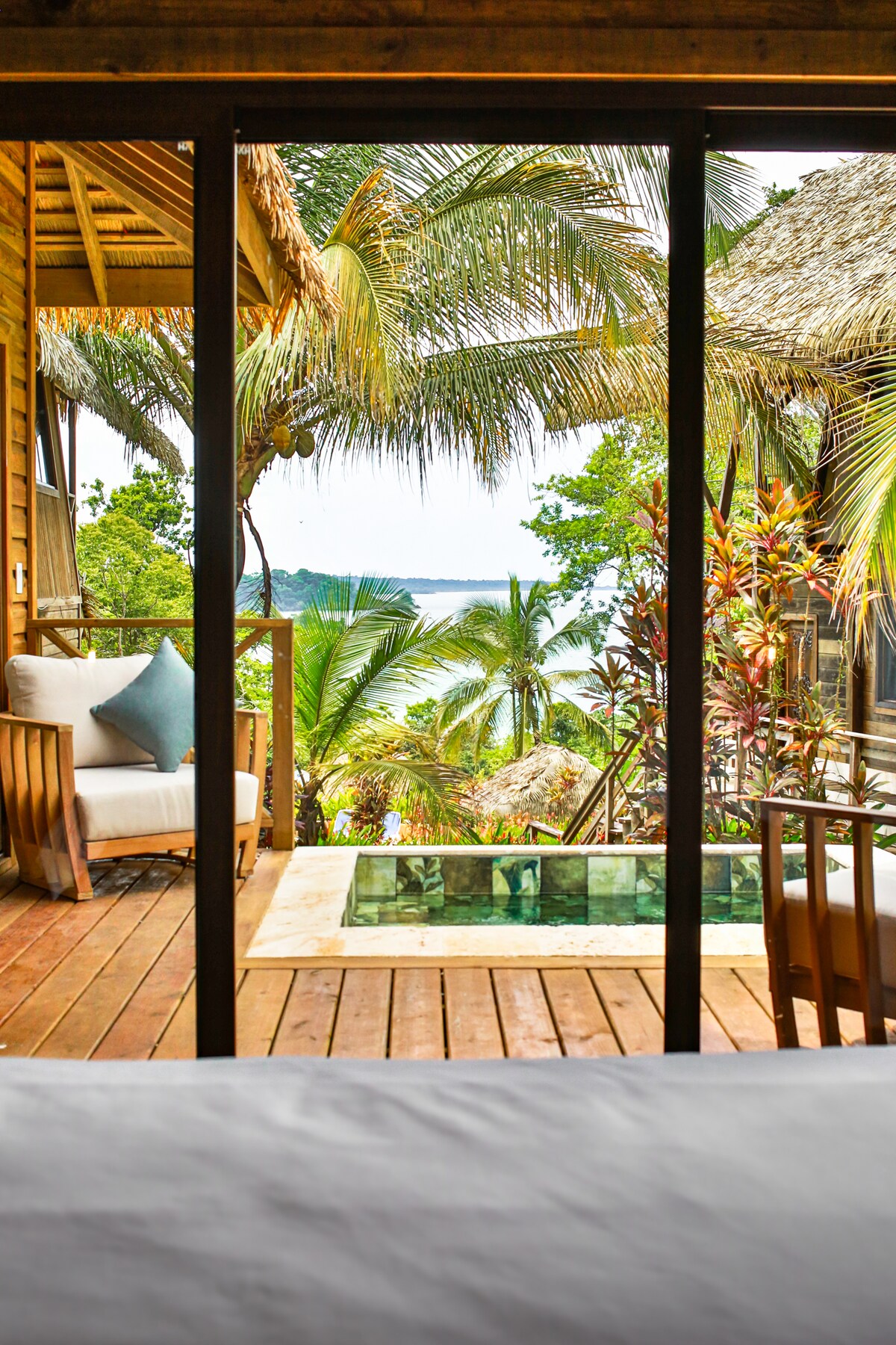 A view from the interior shows sleek glass doors leading to a spacious terrace with a private plunge pool. Lush greenery surrounds the area, along with palm trees that frame an expansive ocean view. A cozy seating arrangement is visible on the terrace.