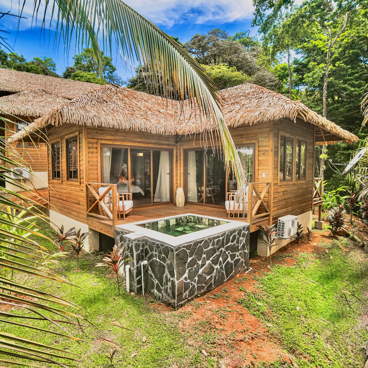 A charming cabin-style retreat nestled among lush greenery features a thatched roof and multiple large windows. A private plunge pool, bordered by stone, can be seen alongside a secluded terrace, ideal for enjoying the natural surroundings.