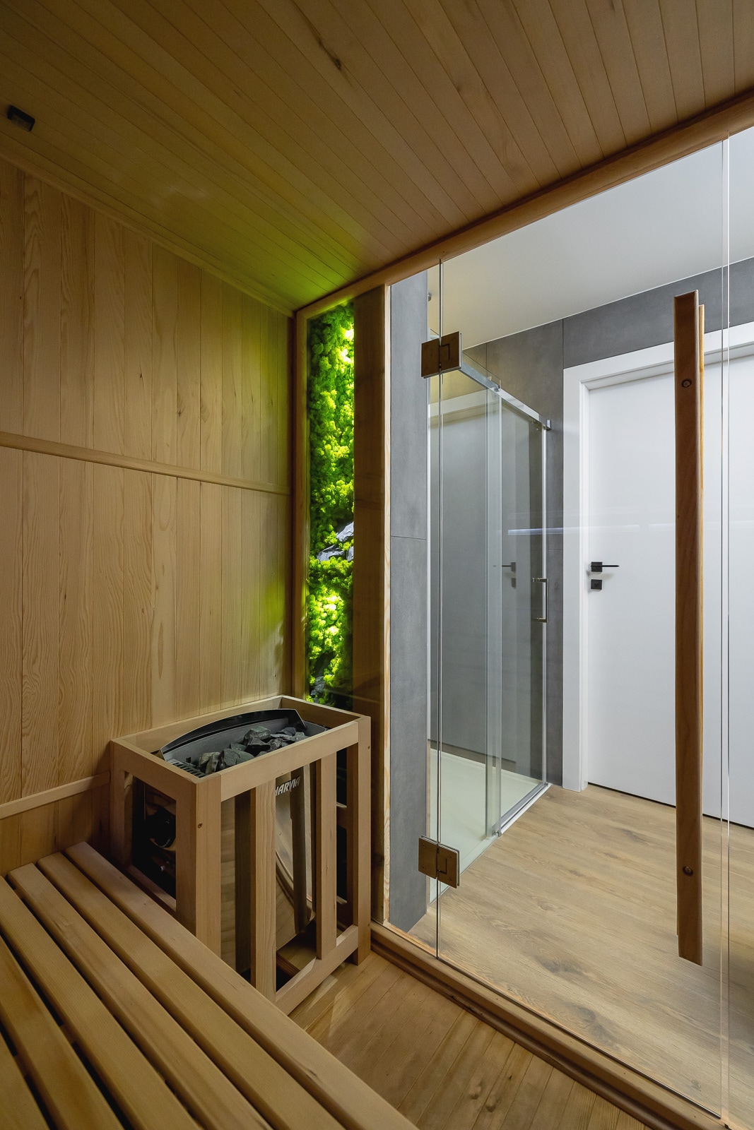 A private sauna room features wooden bench seating and a sleek glass enclosure. A small heater is located beside the seating area, while a portion of the wall is accentuated with greenery, providing a refreshing touch to the space.