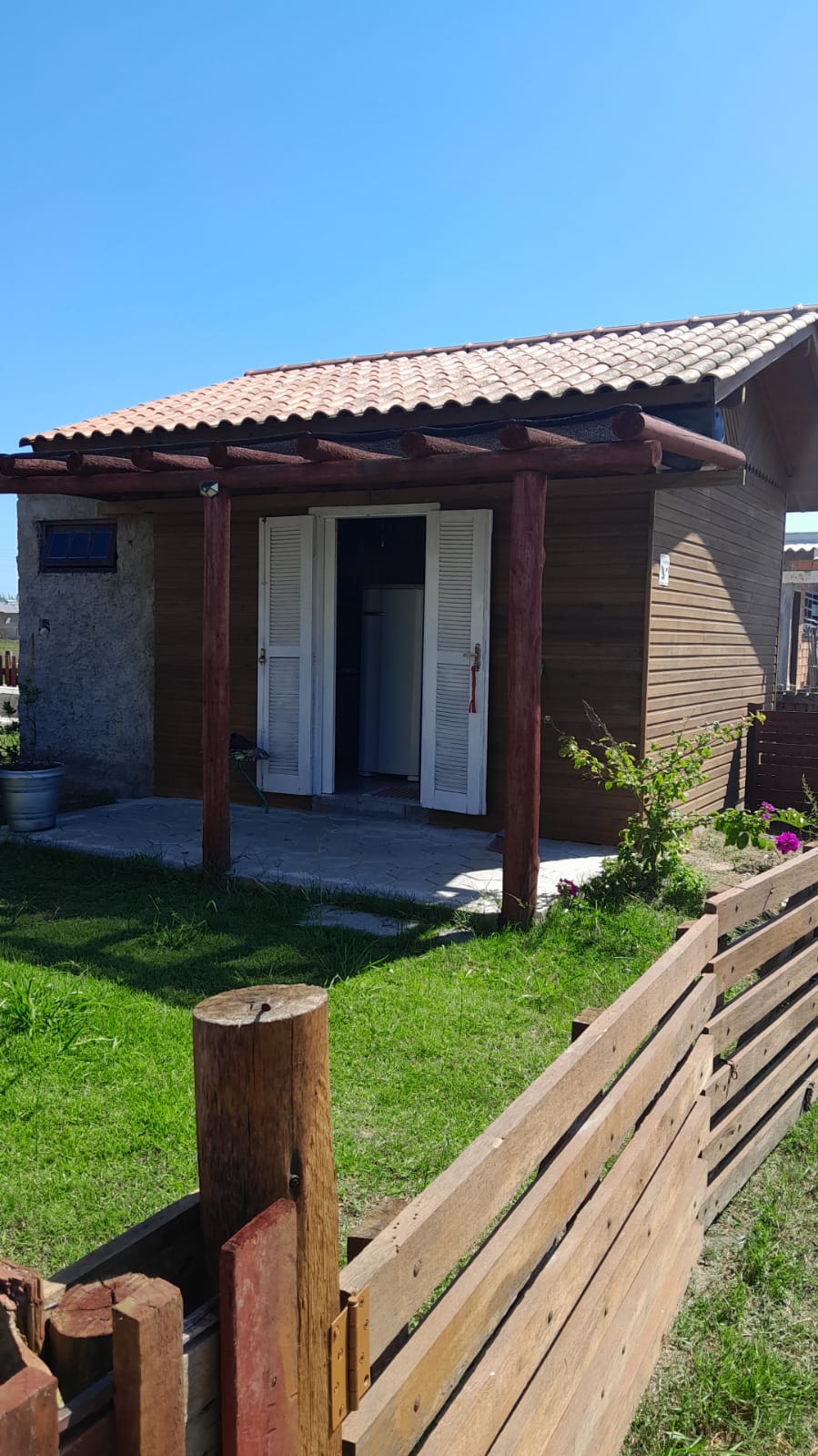 A charming wooden house features a sloped tile roof and an inviting porch area supported by wooden beams. White-shuttered glass doors open to the interior, while a small grassy yard surrounds the entrance, complemented by a wooden fence and scattered flowers.