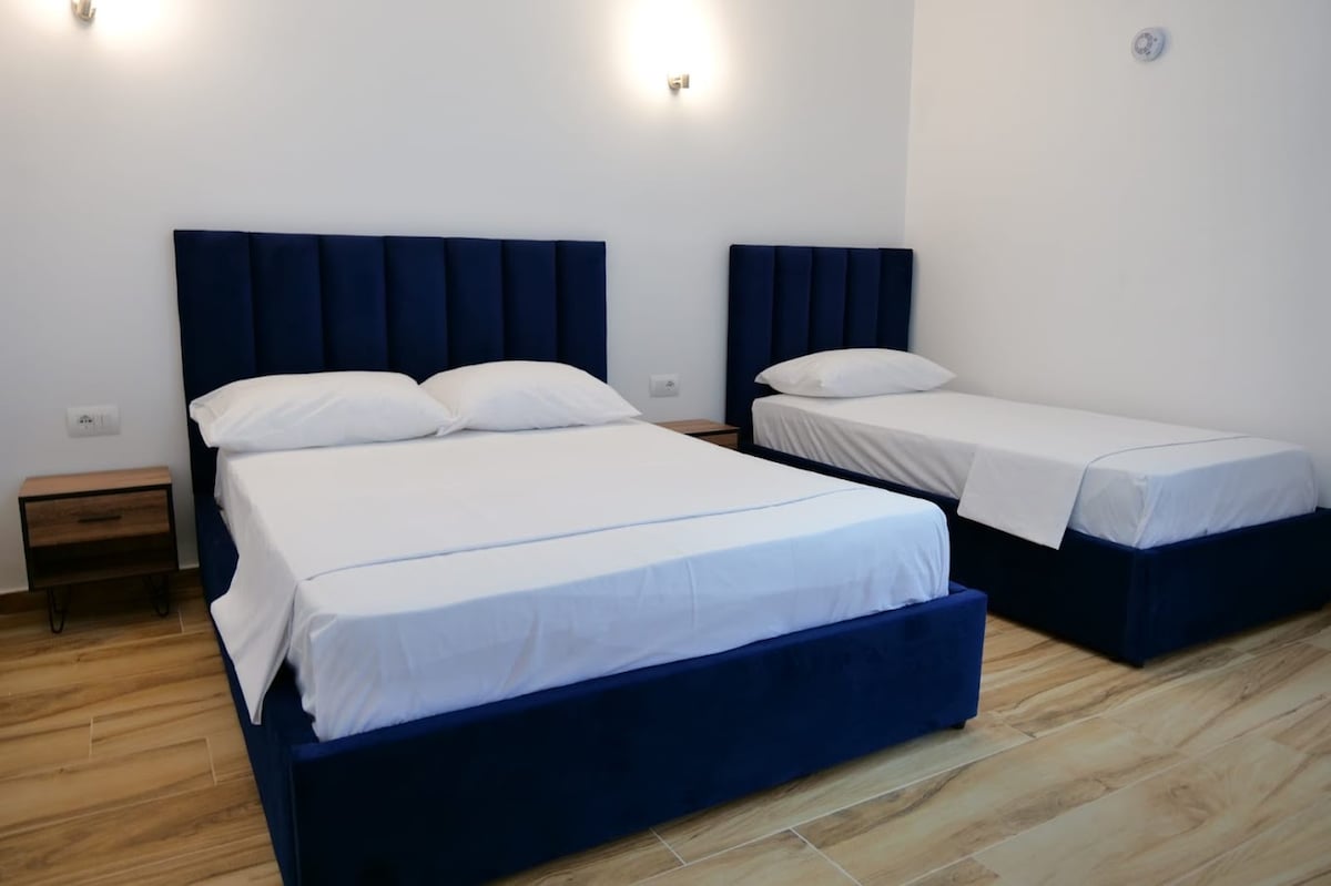 Two stylish beds are positioned in a bright sleeping area, featuring a deep blue upholstered headboard. Nightstands with lamps are placed beside each bed. The floor is covered in light-toned wood, enhancing the spacious feel of the room.