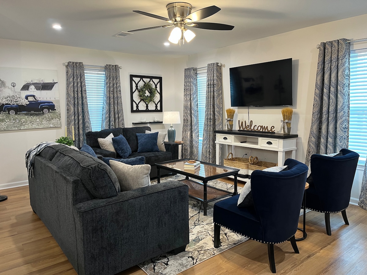 A welcoming living room is highlighted by a large sectional sofa and two accent chairs. Natural light filters through the light blue curtains, illuminating a coffee table at the center. A TV is mounted on the wall, and a side table holds a decorative lamp.