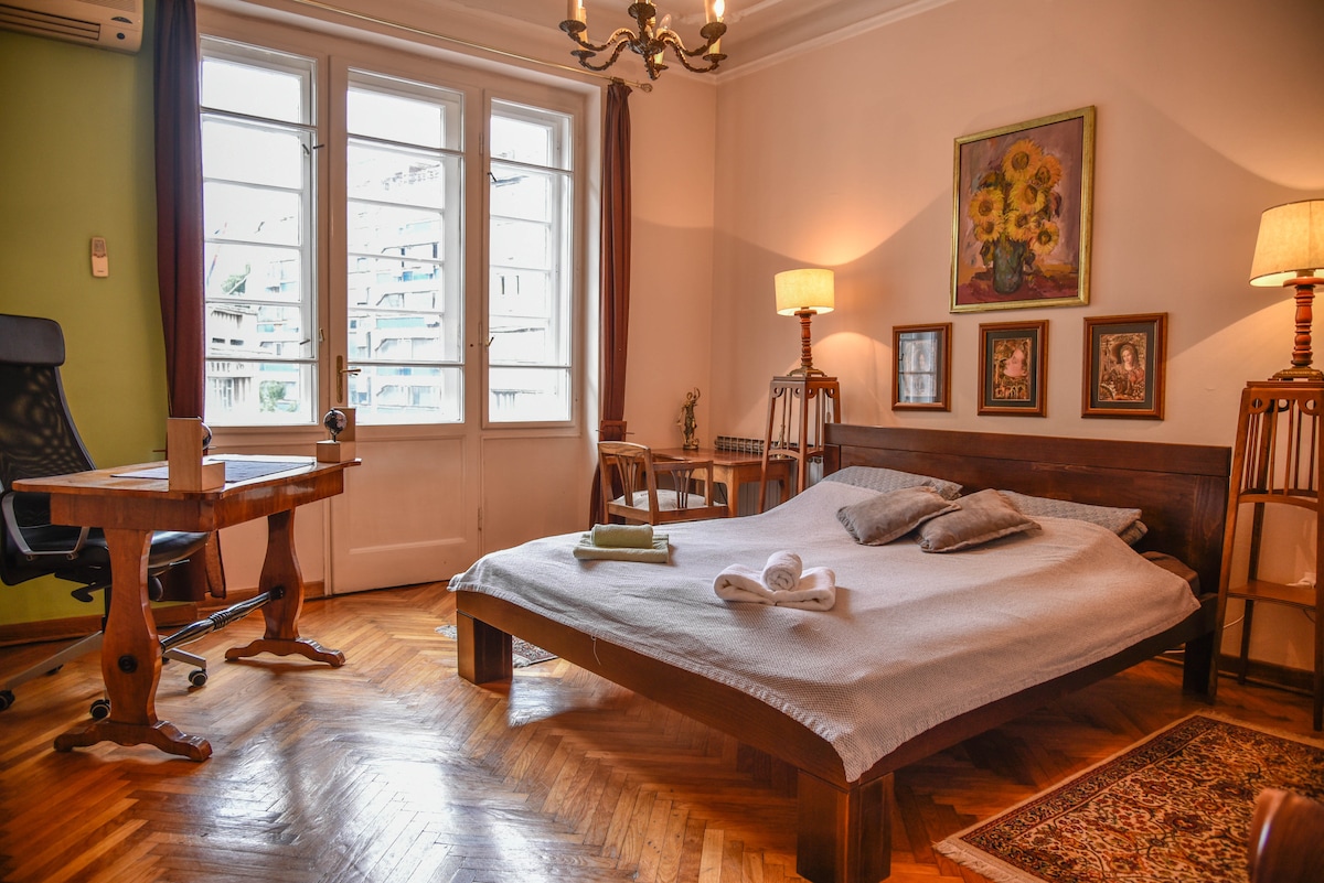 A spacious bedroom features a large bed with light bedding and several pillows. A work desk is positioned nearby, accompanied by an ergonomic chair. Natural light enters through large windows, and decorative lamps provide soft lighting. A colorful painting adorns one wall.