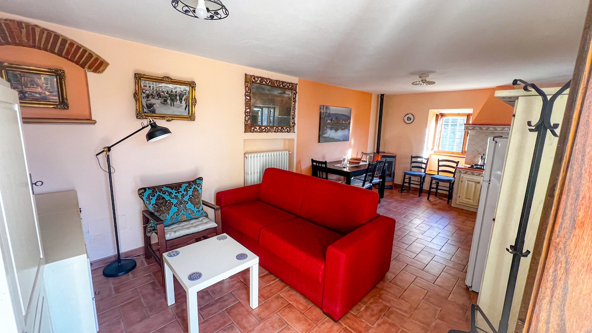 A bright living area features a red sofa and a comfortable chair adorned with a decorative cushion. Artwork lines the walls, complemented by a dining table surrounded by black chairs. A window allows natural light to fill the space, creating a cozy atmosphere.