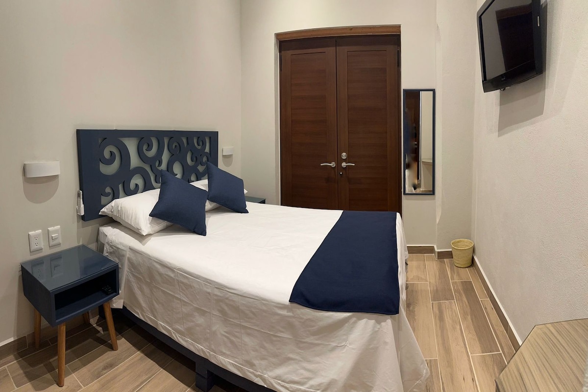 A bedroom features a spacious bed with a navy blue and white bedspread. Two decorative pillows are placed on the bed. A wooden wardrobe with double doors is seen in the background along with a wall-mounted television. A mirror and small bedside table complete the room's layout.