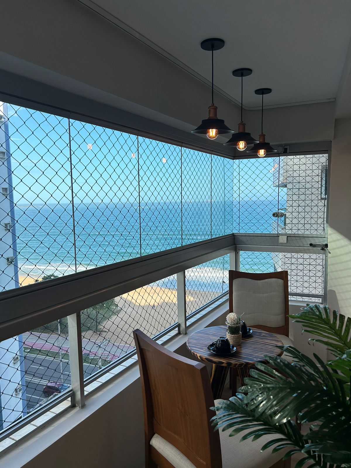A private balcony is featured with a small table and two chairs, positioned near a window showcasing an unobstructed view of the ocean. Decorative pendant lights are suspended above, adding a subtle touch to the serene coastal atmosphere.