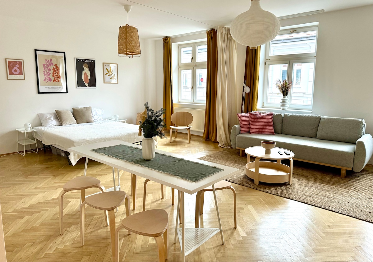 A spacious living area features a light wooden dining table surrounded by four chairs. A cozy sofa is presented beside a window with sheer curtains. A luxurious bed is positioned against the wall, complemented by framed artwork and decorative elements throughout the room.