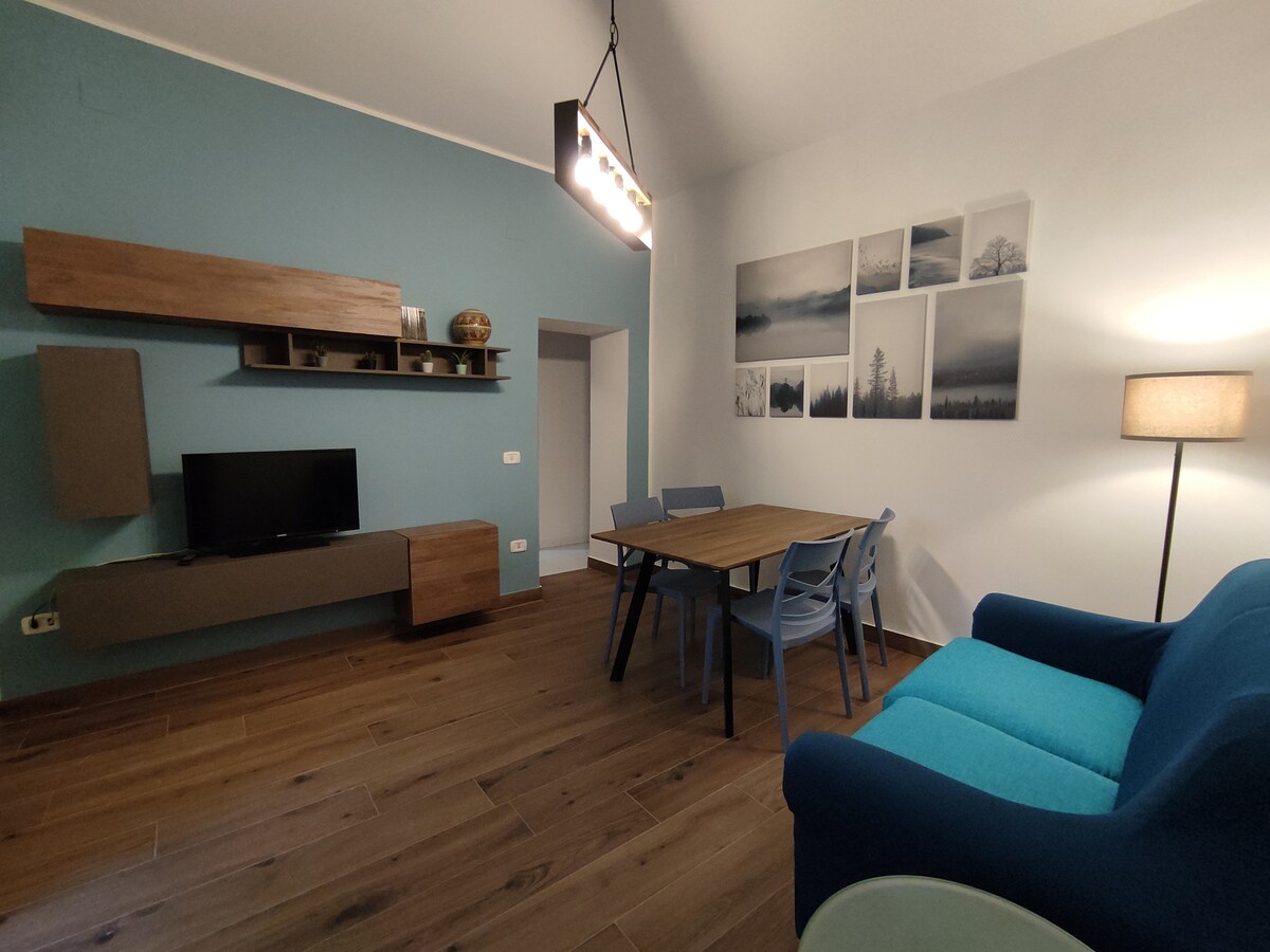 A well-furnished living area features a blue accent wall and wooden flooring. A modern light fixture hangs overhead, illuminating a dining table surrounded by six chairs. A comfortable blue couch complements the space, with a television mounted on a sleek, minimalist media console. Framed landscape images adorn the wall.